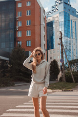 teen walk. young blonde curly girl in sports gray costume is walking in step along the crosswalk and looking with smile on the street buildings background at sunset. lifestyle concept, free space