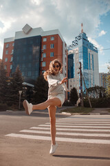 teen walk. young blonde curly girl in sports gray costume is walking with funny jump and smiling happy in sunglasses on the street buildings background. sport lifestyle concept, free space