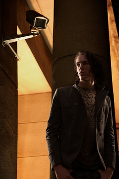 Young man standing outside building at night leaning on column with surveillance camera over his shoulder 