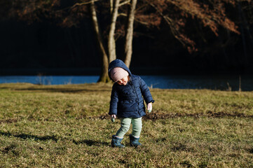 Naklejka na ściany i meble Baby girl in jacket stand in sunny spring park against river.