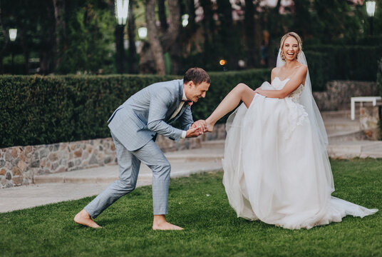 Stylish groom in a gray checkered suit kisses the leg of the blonde bride in a long white dress in a meadow with green grass, against the background of the forest. A funny and funny photo.