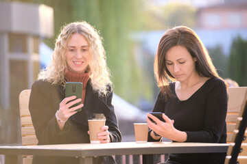 Two distracted women browsing their sellphones sitting at city street cafe. Girlfriends ignoring...