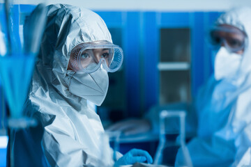 woman in goggles, medical mask and hazmat suit near scientist on blurred background.