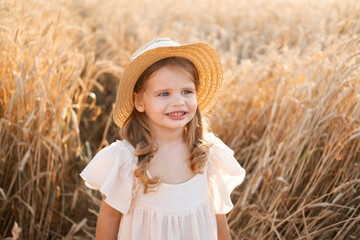 child blonde girl in straw hat and beige muslin dress in wheat field on sunset