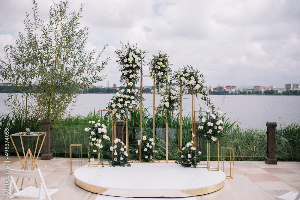 Wall mural Beautiful round wedding arch decorated with flowers and greenery near lake or river outdoors, copy space. Decorations for wedding ceremony in open air