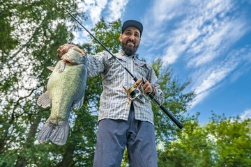 Bass fishing. Big bass fish in hands of pleased fisherman with spinning rod. Largemouth perch at pond. View from bottom - obrazy, fototapety, plakaty