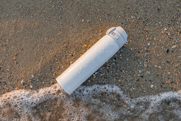 White reusable steel stainless thermo water bottle on sand on sea beach. Flask thermos of drinks...