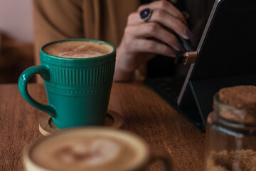 Fototapeta na wymiar Vintage aqua green pastel cappuccino mug with a blurred young woman's hand working on a tablet in the background. Technology, space work, and coffee concept.