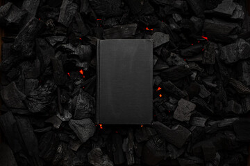 A closed book with a black cover on smoldering charcoal. Textured dark back.