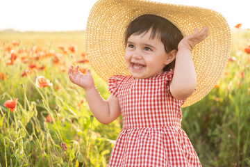 cute baby girl in red dress and straw hat on field of poppies at summer sunset