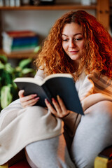 A beautiful ginger girl sitting in the living room wrapped in a blanket and reading an interesting...
