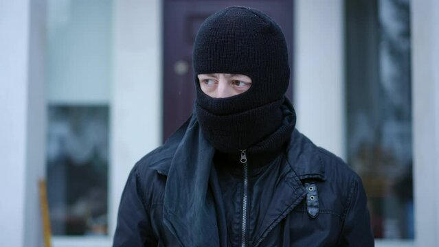 Concentrated robber adjusting balaclava ski mask preparing to steal private property. Portrait of Caucasian man standing outdoors at background of house planning crime