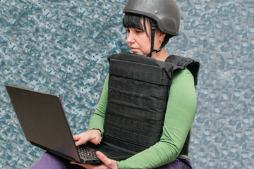 A woman in military uniform is working on a laptop. The girl in the helmet and body armor is a soldier of the information war. Cyber security concept.