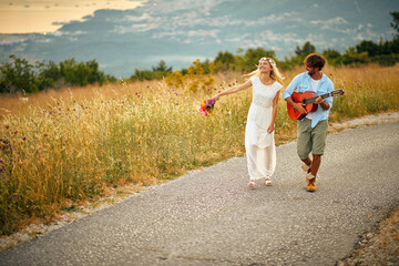 A young groom is playing guitar for his bride while they walking the road in the nature. Relationship, honeymoon, nature