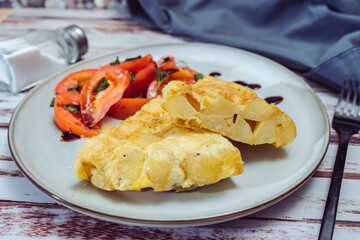high view of a plate with two portions of Spanish potato omelette and sliced tomatoes with olive and sherry vinegar on a wooden table.