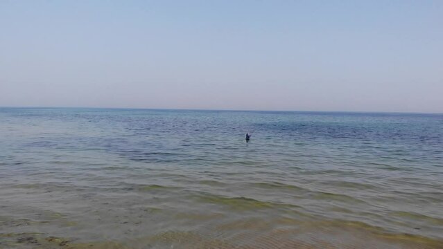 Luftaufnahme von Angler mit Angel in Wathose im Wasser bei Sonne watend per Drohne gefilmt als Fahrt &uuml;ber Strand