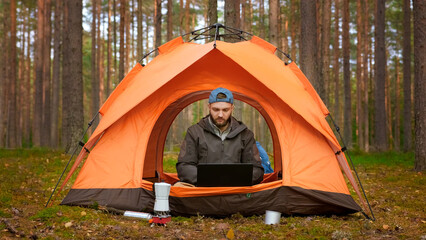 Young man is relaxing in nature sitting in an orange tent, working remotely on laptop, waving his...