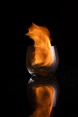 A Single cognac glass on a reflective surface with a black background with a yellow orange flame held within the clear glass
