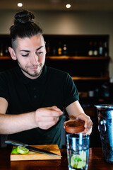 Young, modern waiter, adding brown sugar in a large crystal glass to prepare a Mojito. vertical