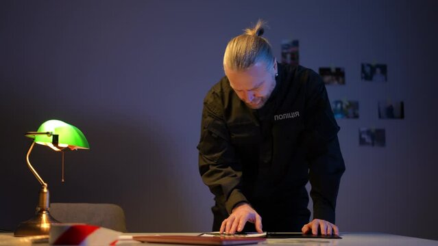 Focused Ukrainian police officer opening case folder examining photos indoors. Portrait of handsome concentrated man in uniform investigating crime in darkness with table lamp lighting