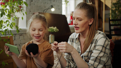 A girl helps her mother to knit. She holds a cell phone in one hand and a ball of nits in the other - obrazy, fototapety, plakaty