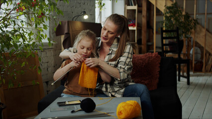 A woman helps a girl learn to knit. She hugs her and holds her hands