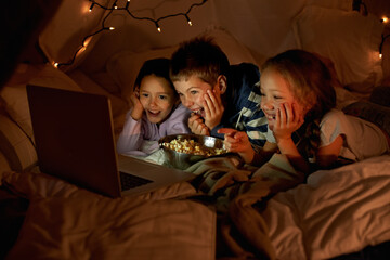 Kids are never far from technology. Shot of three young children using a laptop in a blanket fort. - Powered by Adobe