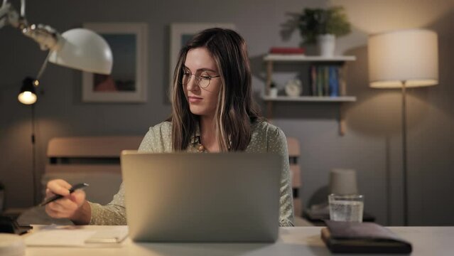 Freelancer works from home. Focused woman is using computer, typing text and making notes on paper. Student studying doing tasks on laptop
