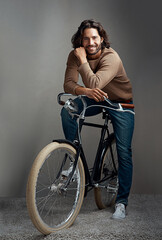 Obraz premium The hippest way to get around. Studio shot of a handsome young man leaning against a bicycle in front of a gray background.