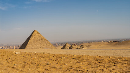 Fototapeta na wymiar Ancient Egyptian pyramids: a large Menkaura and three small pyramids of queens nearby. In the foreground is the yellow sand of the desert. In the distance- silhouettes of buildings in Cairo. Blue sky.