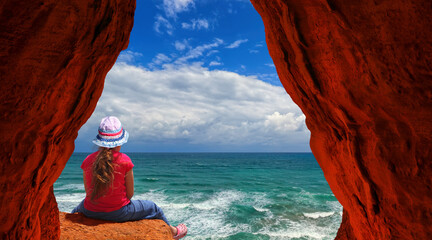 Little girl in sun hat sitting on a stone among the red rocks. She looks from high at the sea 