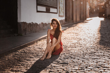 Sexy young Caucasian girl in a red dress posing sitting on a paving stone on a beautiful European...