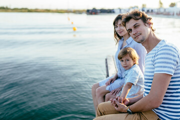 Fototapeta na wymiar Family sitting at the edge of dock over lake. Side view of dad, mom little son family outdoor recreation.