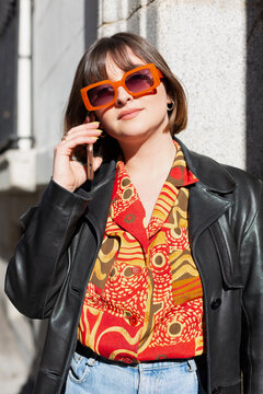 Portrait of young fashionable caucasian woman talking on the phone on the street. She is wearing a shirt with orange shades and matching sunglasses. Cool style and female fashion concept. 