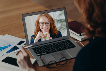 Fototapeta na wymiar Female freelancer talks with employee by video call, discuss strategy of work. Laptop screen view of smiling ginger woman in spectacles