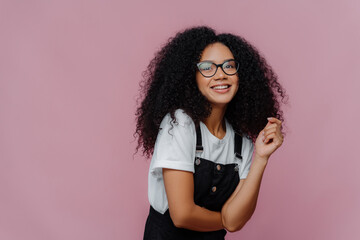Happy woman with crisp hair, has dreamy expression, wears optical glasses, stylish clothes stands...