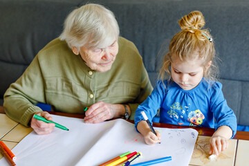 Beautiful toddler girl and grand grandmother drawing together pictures with felt pens at home. Cute...