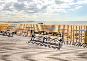 Fototapeta na wymiar Benches at Coney island Beach Promenade, rear view, with Ocean and Pat Auletta Steeplechase Pier in the background, New York City during sunny winter day with cloudy sky, horizontal