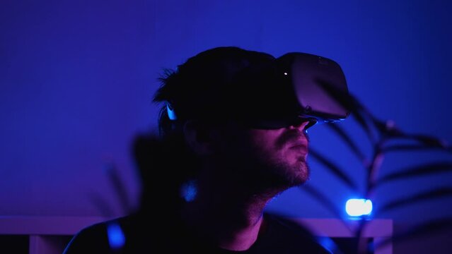 Close-up of a man wearing VR glasses in a dark room with blue light spinning his head. There is a room flower in the front captivity. VR helmet for entertainment, learning and gaming
