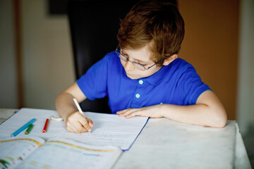 Portrait of little kid boy with glasses at home making homework, writing letters with colorful pens. Little child doing exercise, indoors. Elementary school and education, home schooling concept.