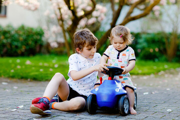 Cute little baby girl and school kid boy playing with blue small toy car in garden of home or...