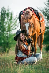 Foto op Canvas A cowgirl sits on the ground near her horse in a field in summer. © sergo321