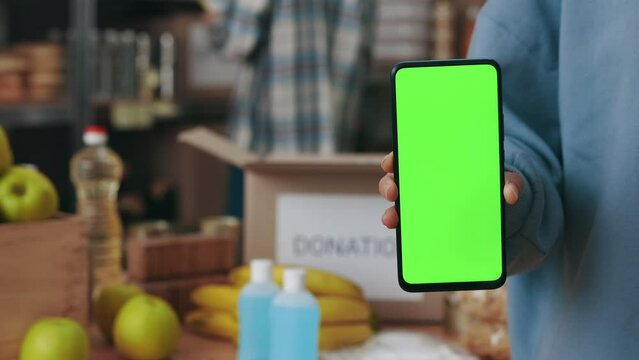 Close up of woman holding modern smartphone with chroma key screen while man on background packing donation boxes. Technology, charity and people concept.