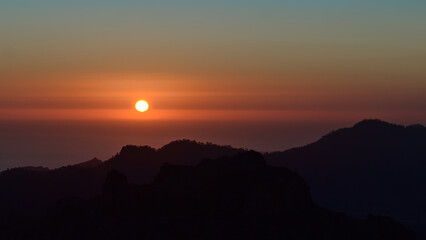 Stunning sunset above the rugged mountains of island Gran Canaria, Canary Islands, Spain with red sun in the colorful sky and the silhouettes of hills.