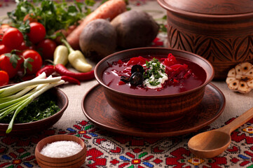 Red Ukrainian borscht in a beautiful brown ceramic dish. There are a lot of fresh vegetables nearby: carrots, beets, garlic, herbs, tomatoes