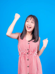 Portrait of excited Beautiful Asian woman cute girl in red dress with bangs hair style raising hand in the air laughing cheerfully gesturing for winner , smiling , look camera isolated blue background