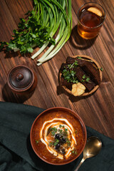 On a dark wooden table there is a clay plate with red Ukrainian borsch, next to croutons with garlic and fresh herbs - onion and parsley