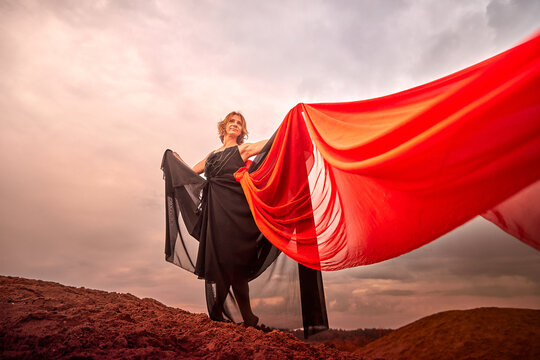 A girl in black dress with red fabric dances on sand dunes against a dramatic sky before a thunderstorm. Model posing during photo shoot on nature