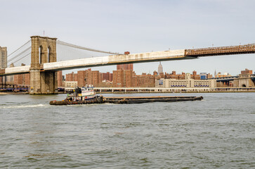 Industrial Boat pushes a flat platform on east river underneath Brooklyn Bridge with construction area on the bridge, New York City, horizontal