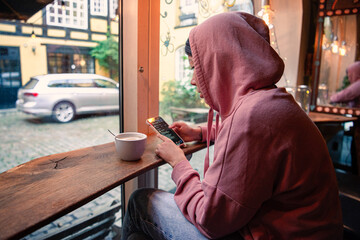 A girl with black hair in a hoodie and jeans looks into her smartphone and drinks a cappuccino coffee while sitting near the window in a cafe. Copenhagen, Denmark